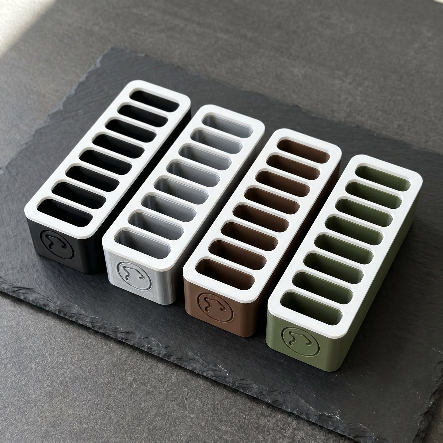 Norello Fuel Rack in vier Farben nebeneinander: Nachtschwarz, Grau, Braun, Olivgrün — je 8 Slots, weißer Deckel